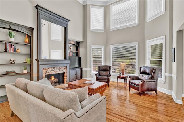 living room with crown molding, a high ceiling, light wood-style floors, a stone fireplace, and baseboards