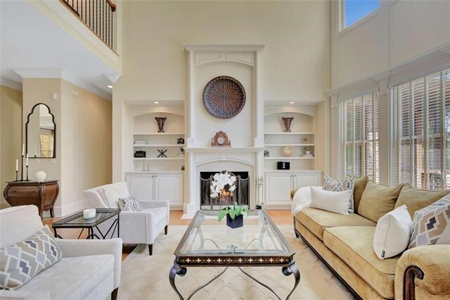 living room with a high ceiling, built in features, a lit fireplace, ornamental molding, and baseboards