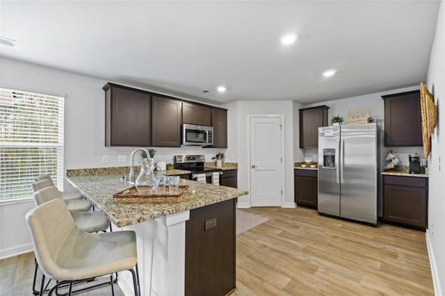 kitchen with a breakfast bar area, dark brown cabinets, light hardwood / wood-style flooring, appliances with stainless steel finishes, and light stone countertops