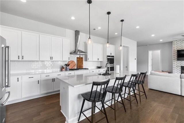 kitchen featuring wall chimney exhaust hood, a kitchen island with sink, hanging light fixtures, and dark hardwood / wood-style flooring