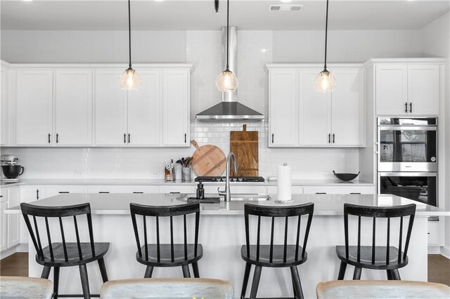 kitchen with hanging light fixtures, double oven, a center island with sink, and white cabinets