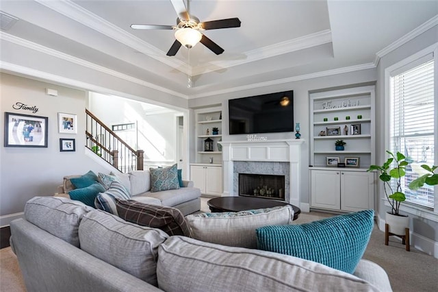 carpeted living room featuring built in shelves, ceiling fan, crown molding, and a raised ceiling