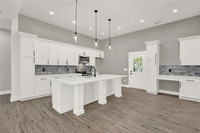 kitchen with appliances with stainless steel finishes, decorative light fixtures, white cabinetry, wood-type flooring, and a kitchen island with sink