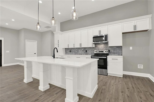 kitchen with sink, appliances with stainless steel finishes, a kitchen island with sink, white cabinets, and decorative light fixtures