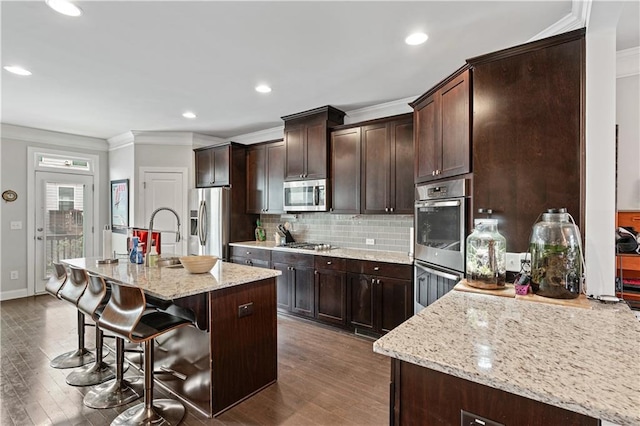 kitchen featuring dark wood-type flooring, a breakfast bar area, appliances with stainless steel finishes, light stone countertops, and an island with sink