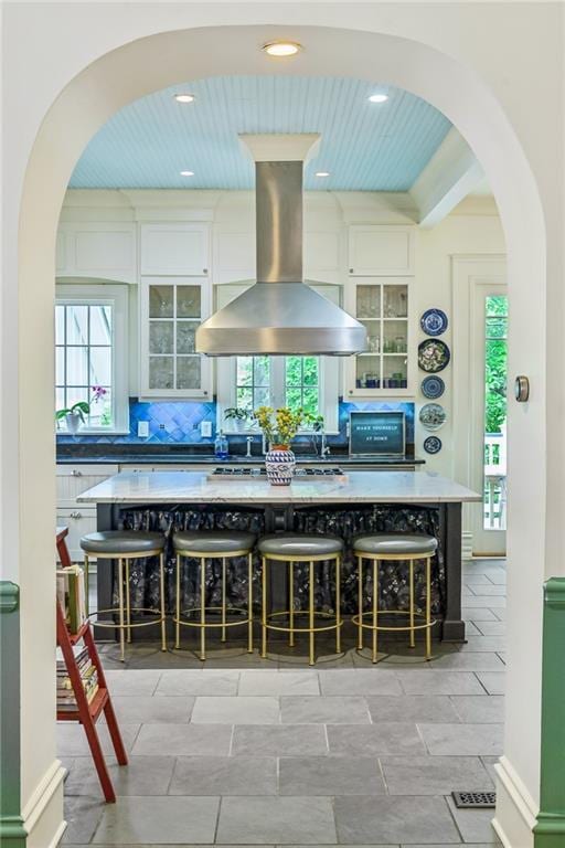 interior space featuring island exhaust hood, arched walkways, tasteful backsplash, glass insert cabinets, and white cabinetry