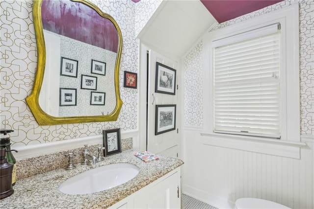 half bathroom featuring vanity, a wainscoted wall, lofted ceiling, a decorative wall, and wallpapered walls
