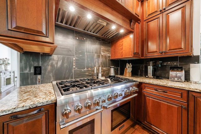 kitchen featuring double oven range, premium range hood, tasteful backsplash, light stone countertops, and brown cabinets