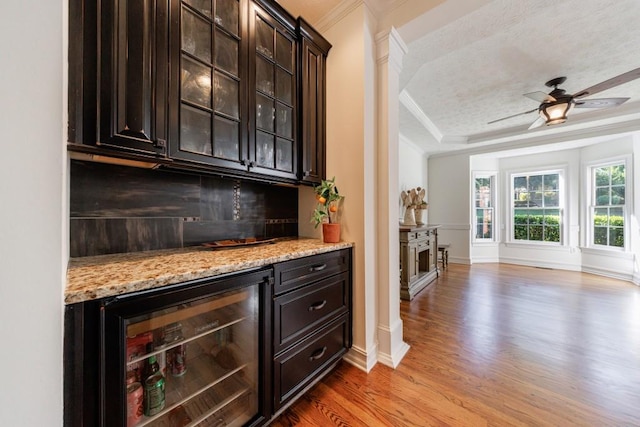 indoor dry bar featuring beverage cooler, crown molding, light wood-style floors, decorative backsplash, and a tray ceiling