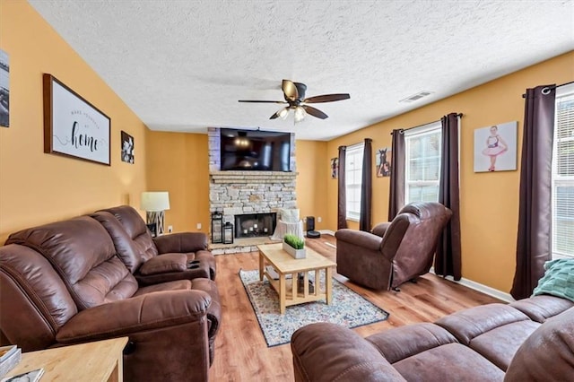 living room featuring a textured ceiling, light hardwood / wood-style flooring, a stone fireplace, and ceiling fan