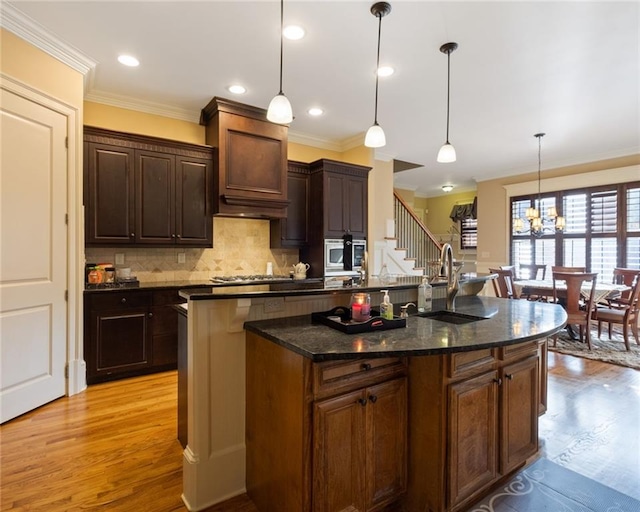 kitchen with sink, light hardwood / wood-style flooring, dark stone countertops, an island with sink, and decorative light fixtures