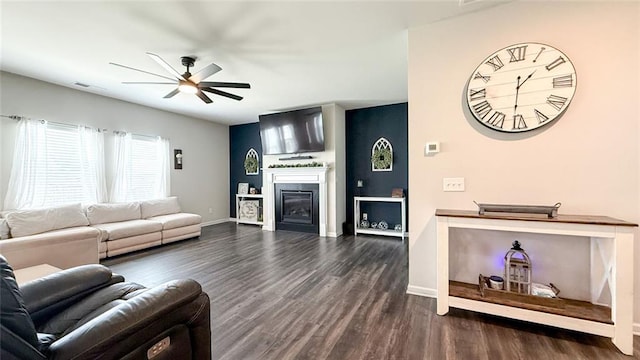 living room with a ceiling fan, visible vents, baseboards, a glass covered fireplace, and dark wood-type flooring
