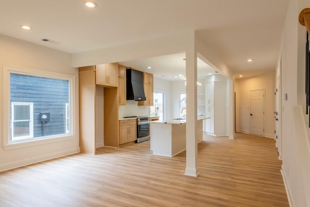 kitchen featuring wall chimney exhaust hood, light brown cabinetry, range with two ovens, light hardwood / wood-style floors, and decorative backsplash