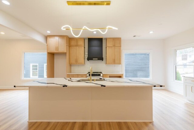 kitchen featuring a kitchen island with sink, wall chimney range hood, decorative light fixtures, and light brown cabinetry