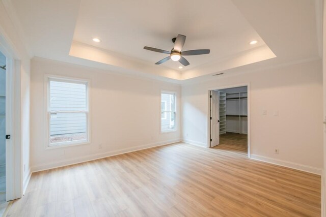 unfurnished bedroom with ceiling fan, a tray ceiling, a spacious closet, and light wood-type flooring