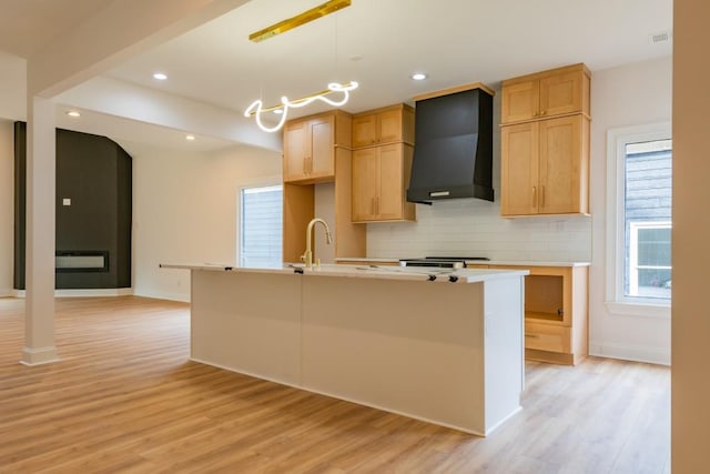 kitchen with light hardwood / wood-style flooring, a center island with sink, decorative light fixtures, wall chimney exhaust hood, and light brown cabinets