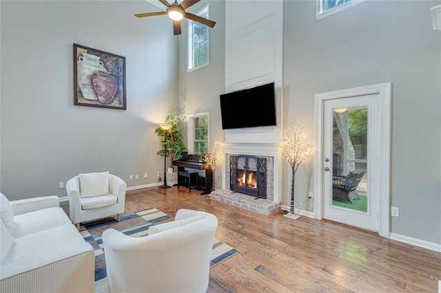 living room featuring a towering ceiling, wood finished floors, ceiling fan, a brick fireplace, and baseboards