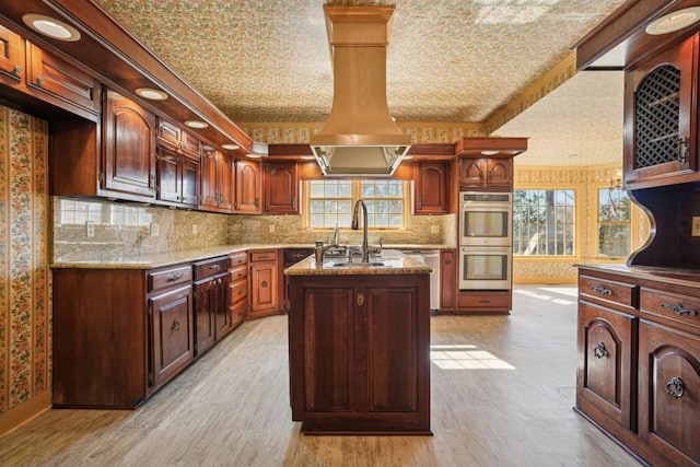 kitchen with wallpapered walls, light wood-type flooring, an island with sink, appliances with stainless steel finishes, and a sink