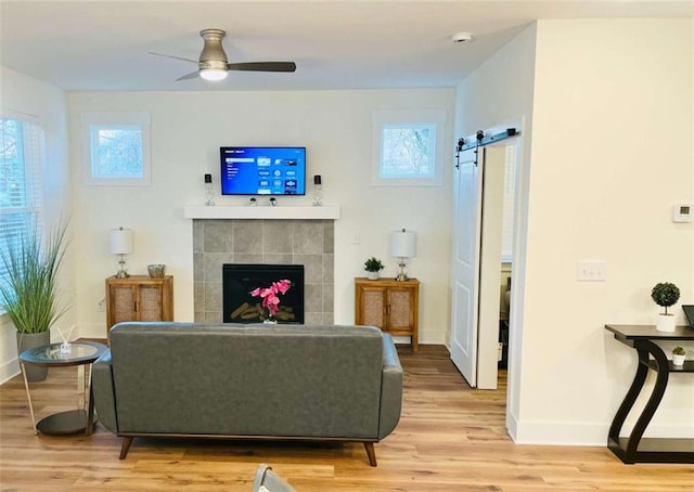 living room with a barn door, a tile fireplace, a ceiling fan, baseboards, and light wood-style floors