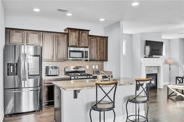 kitchen featuring light stone countertops, appliances with stainless steel finishes, dark brown cabinetry, dark wood-type flooring, and a tiled fireplace