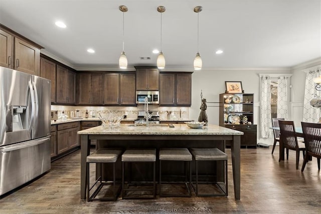 kitchen with stainless steel appliances, light stone counters, pendant lighting, dark brown cabinetry, and a kitchen island with sink