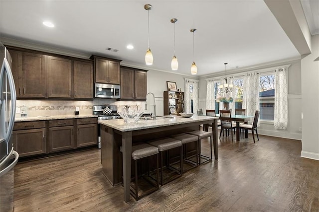 kitchen featuring stainless steel appliances, decorative light fixtures, light stone countertops, a center island with sink, and crown molding