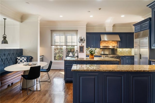 kitchen with a kitchen island with sink, stainless steel appliances, blue cabinetry, wall chimney range hood, and dark wood finished floors