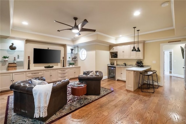 living area with beverage cooler, a ceiling fan, light wood-style flooring, a tray ceiling, and crown molding