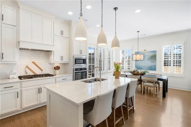 kitchen featuring sink, a kitchen island with sink, stainless steel appliances, white cabinets, and decorative light fixtures