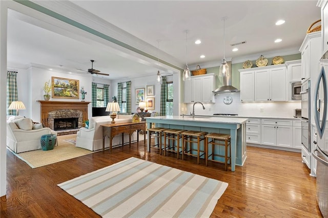 kitchen with wall chimney exhaust hood, hanging light fixtures, a center island with sink, a kitchen bar, and white cabinets