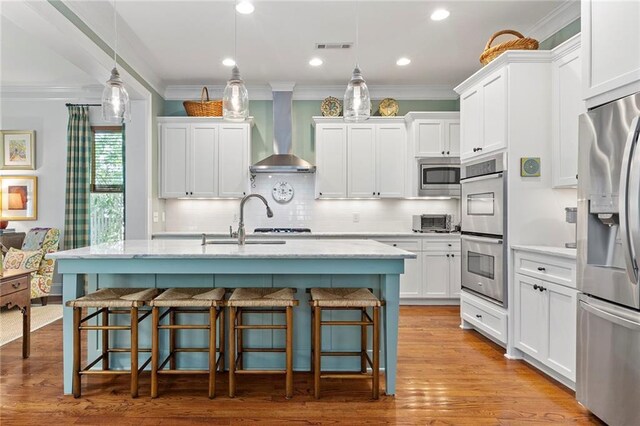 kitchen with stainless steel appliances, decorative light fixtures, white cabinetry, wall chimney exhaust hood, and an island with sink