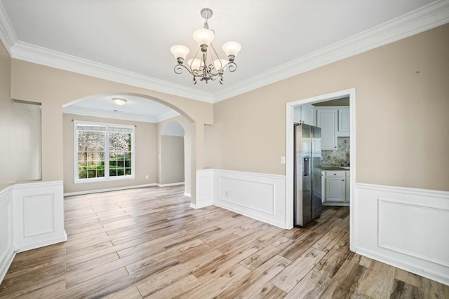 unfurnished dining area with a wainscoted wall, a notable chandelier, light wood-style flooring, arched walkways, and crown molding