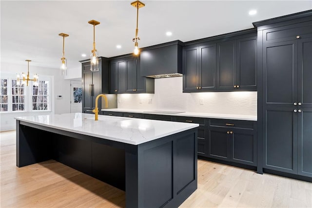 kitchen with an island with sink, sink, hanging light fixtures, wall chimney range hood, and light hardwood / wood-style flooring