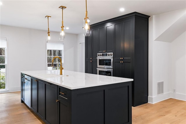 kitchen with sink, appliances with stainless steel finishes, light stone countertops, a center island with sink, and decorative light fixtures