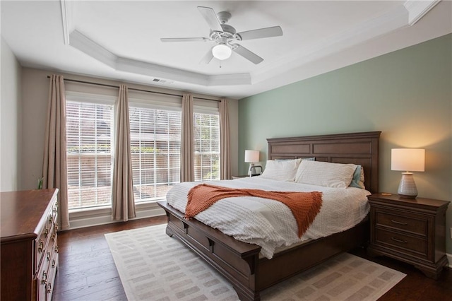 bedroom with visible vents, ceiling fan, a tray ceiling, ornamental molding, and dark wood-style flooring