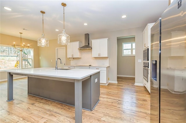 kitchen featuring stainless steel appliances, wall chimney range hood, a center island with sink, white cabinets, and hanging light fixtures