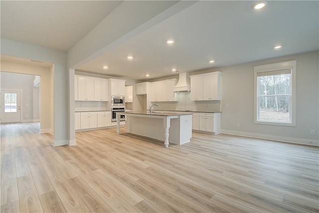 kitchen featuring a center island with sink, custom exhaust hood, stainless steel appliances, and light hardwood / wood-style flooring