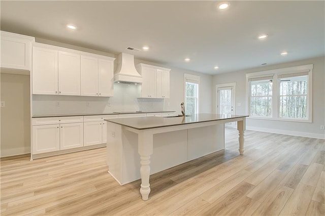 kitchen with light wood-type flooring, custom exhaust hood, a kitchen island with sink, sink, and white cabinetry