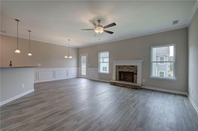 unfurnished living room with dark wood-type flooring, a stone fireplace, ceiling fan with notable chandelier, and crown molding