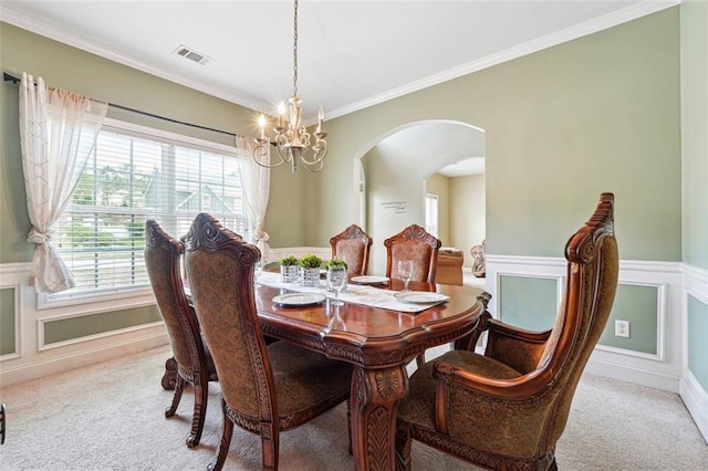 carpeted dining room with a chandelier, arched walkways, crown molding, and wainscoting
