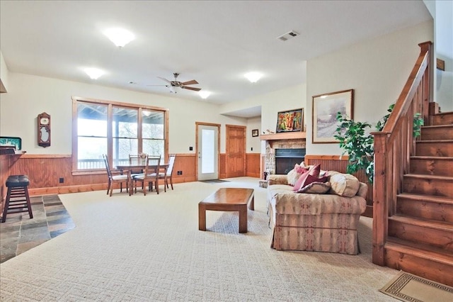 living room with ceiling fan, a stone fireplace, light carpet, and wooden walls