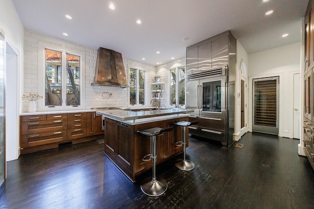 kitchen with a kitchen breakfast bar, wall chimney range hood, tasteful backsplash, dark wood finished floors, and a center island with sink