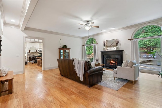 living room featuring a glass covered fireplace, ceiling fan, light wood-style floors, crown molding, and baseboards