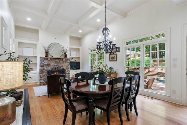 dining area featuring light wood-style floors, a stone fireplace, coffered ceiling, a chandelier, and beam ceiling