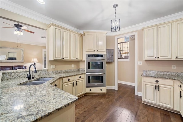 kitchen with cream cabinetry, dark wood finished floors, a sink, double oven, and ceiling fan with notable chandelier