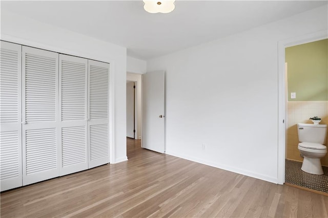 unfurnished bedroom with a wainscoted wall, tile walls, light wood finished floors, a closet, and ensuite bathroom