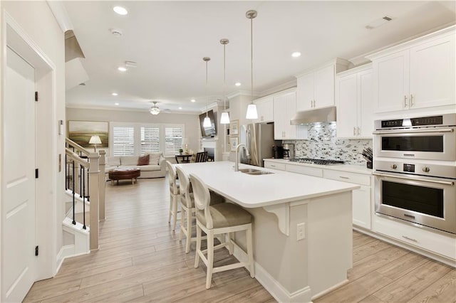 kitchen with under cabinet range hood, stainless steel appliances, open floor plan, decorative backsplash, and an island with sink