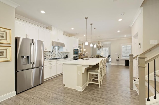 kitchen with under cabinet range hood, stainless steel appliances, a sink, an island with sink, and crown molding