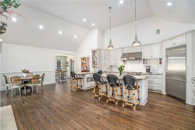 kitchen with under cabinet range hood, a breakfast bar area, stainless steel built in fridge, dark wood-style floors, and open shelves