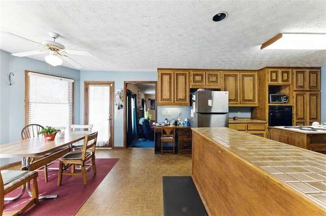 kitchen featuring tile counters, brown cabinetry, freestanding refrigerator, a textured ceiling, and black oven